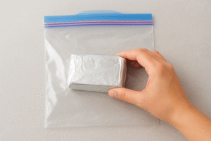 A block of cream cheese in its original packaging being placed inside a freezer-safe bag, illustrating proper double-wrapping for freezer burn prevention.