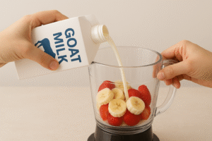 A pair of hands pouring goat milk into a blender along with fruits, illustrating the preparation of a healthy morning smoothie.