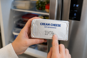 Hand checking the printed expiration date on a foil-wrapped block of fresh cream cheese.