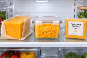 Unopened cheddar cheese block and shredded cheddar in a glass container stored in a clean refrigerator